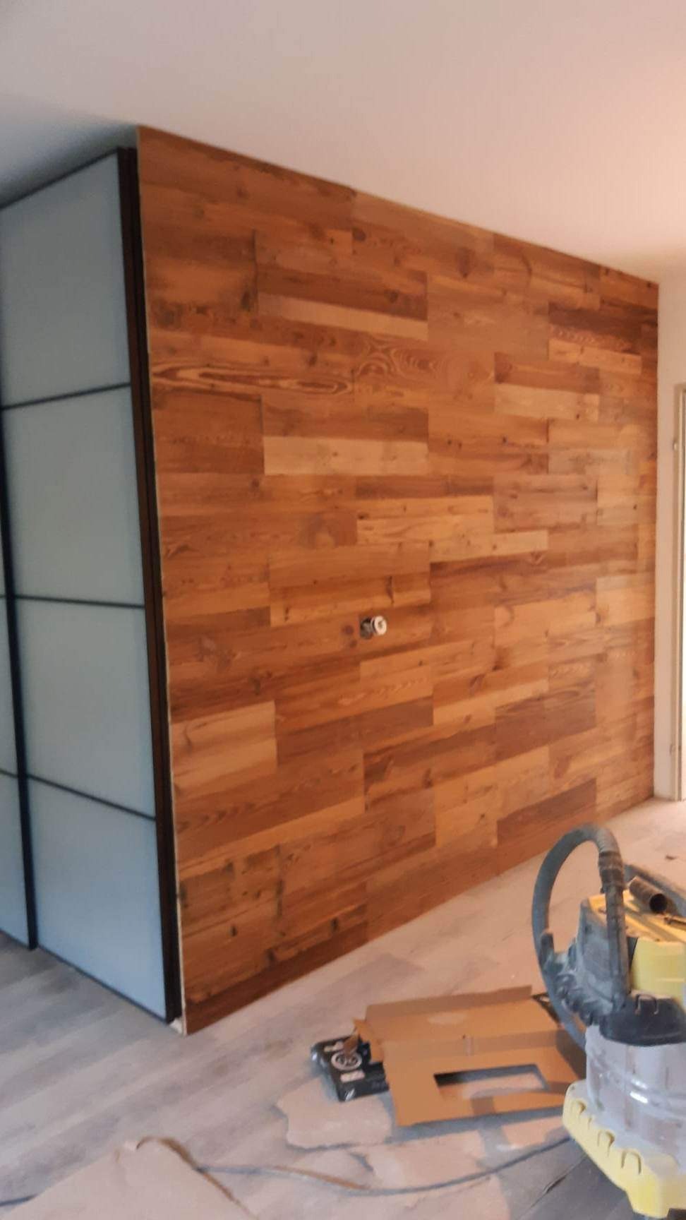 Ściana wykończona panelami z naturalnego drewna o ciepłym odcieniu, z widocznymi gniazdkami elektrycznymi i fragmentem szafy z przesuwnymi drzwiami po lewej stronie. Na podłodze widoczny sprzęt...