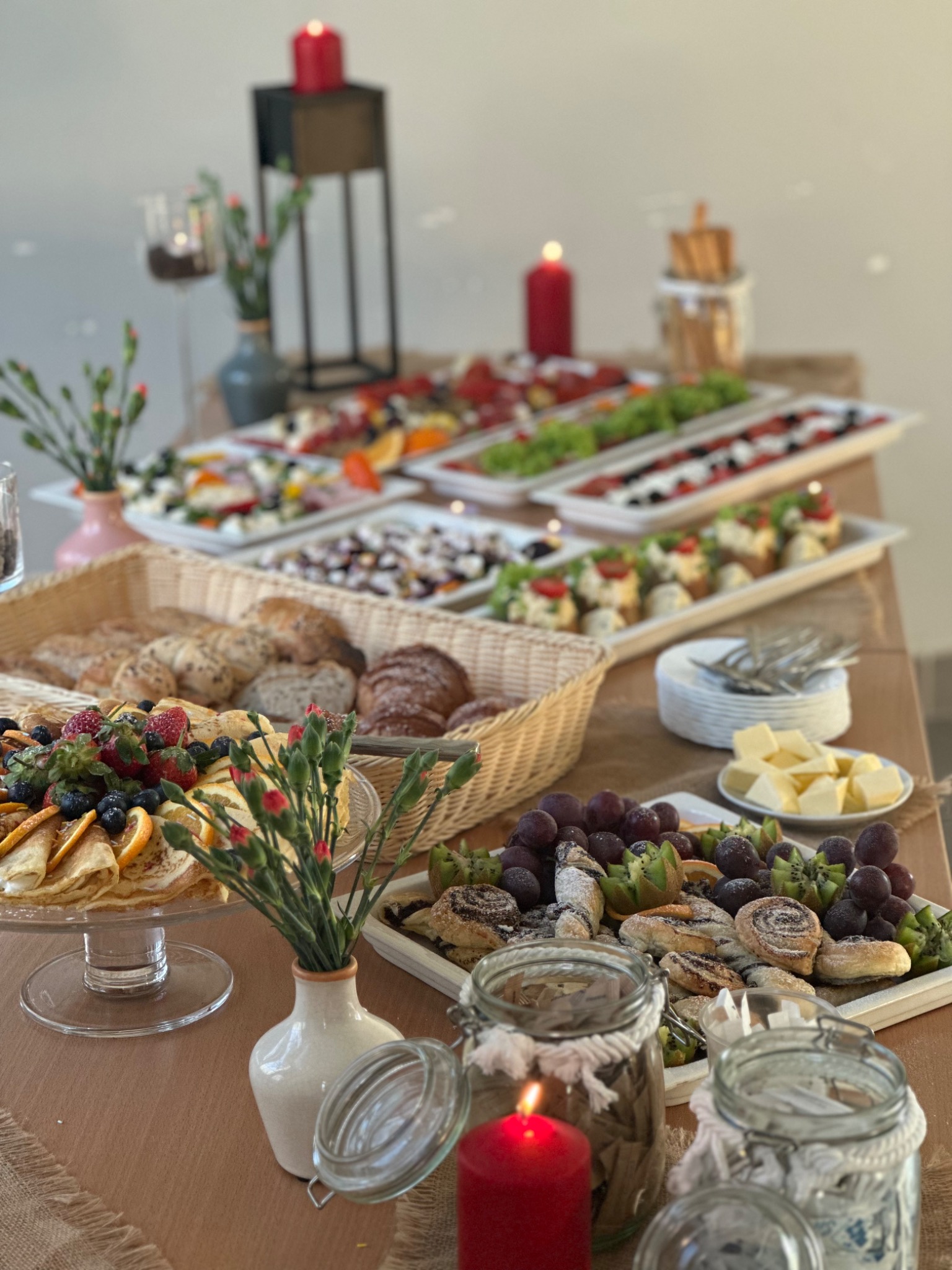 Stół z różnorodnym jedzeniem, w tym pieczywem w koszyku, talerzem naleśników z owocami, sałatkami na tacach, ciastkami, winogronami i kiwi, udekorowany świecami i kwiatami w wazonach.