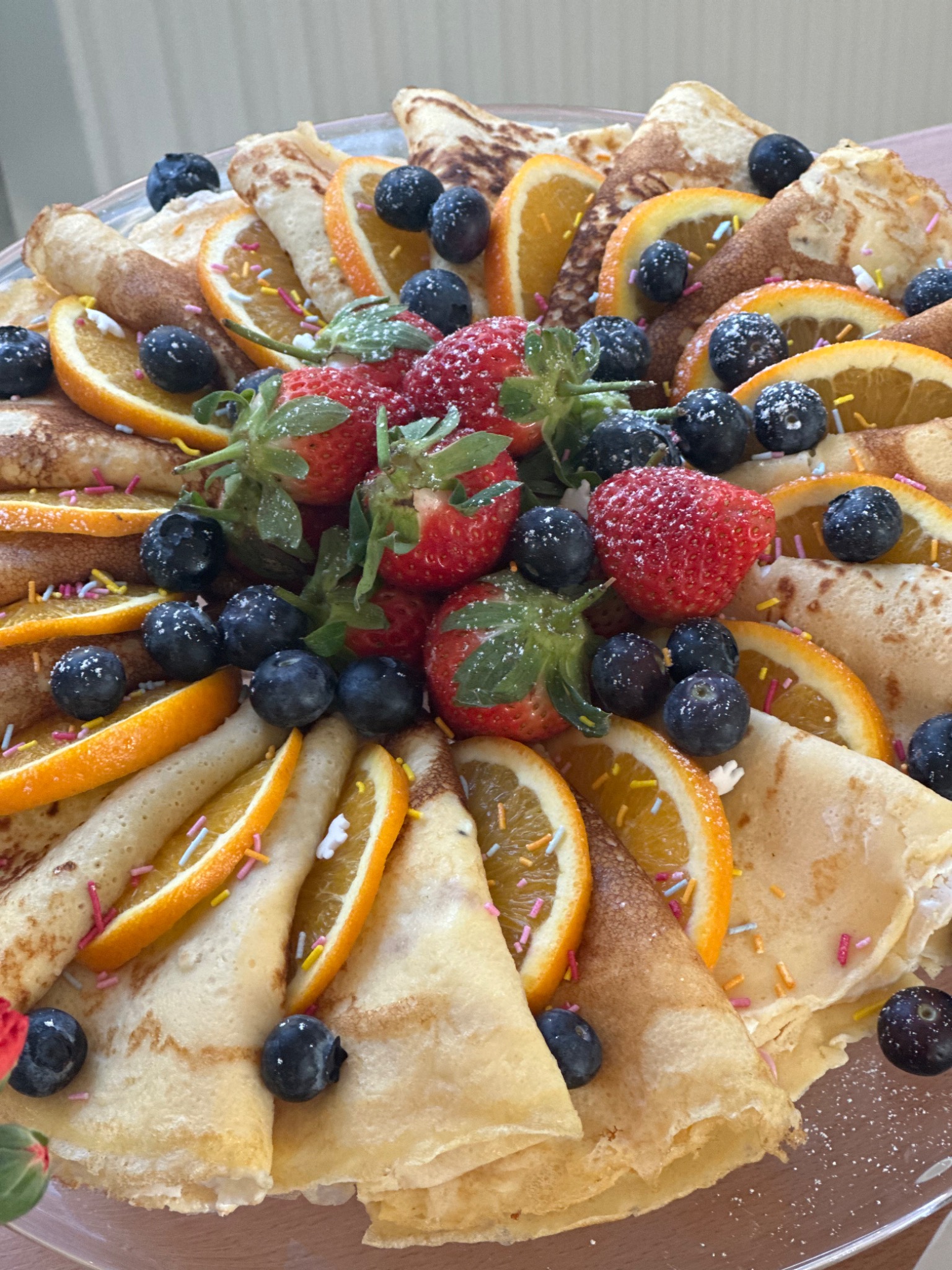 Dekoracyjny talerz z naleśnikami, pomarańczami, borówkami i truskawkami, posypany kolorową posypką i cukrem pudrem, widok z góry.