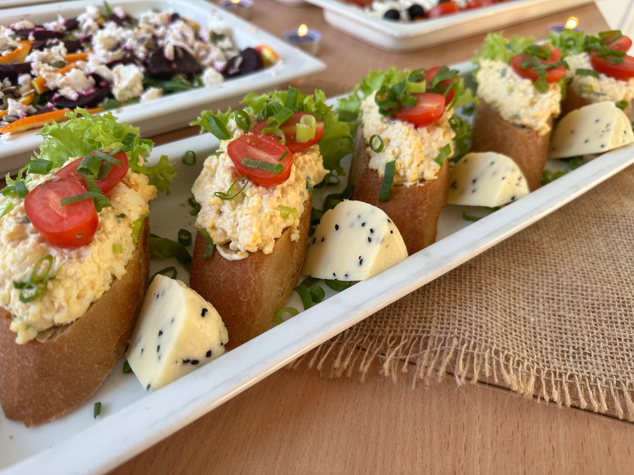 Elegancko podane kanapeczki na białej prostokątnej tacy, z pastą jajeczną, sałatą, pomidorkami cherry i szczypiorkiem, obok ser z czarnuszką, na tle lnianej serwety i innych dań.