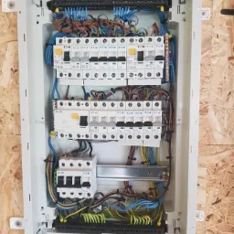 Otwarta skrzynka rozdzielcza z zamontowanymi bezpiecznikami i okablowaniem, umieszczona na ścianie wykończonej płytą OSB. Widoczne rurki karbowane doprowadzające kable.