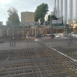 Ernest Sobaszek - Zbrojenie fundamentu z widocznymi prętami stalowymi, szalunkiem i pracownikami w odblaskowych kamizelkach na placu budowy, w tle budynki i silosy.