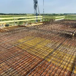 Zbrojenie płyty fundamentowej z widocznymi prętami stalowymi ułożonymi w siatkę na żółtych deskach szalunkowych, w tle krajobraz z zielonymi polami i lasem oraz żuraw budowlany.