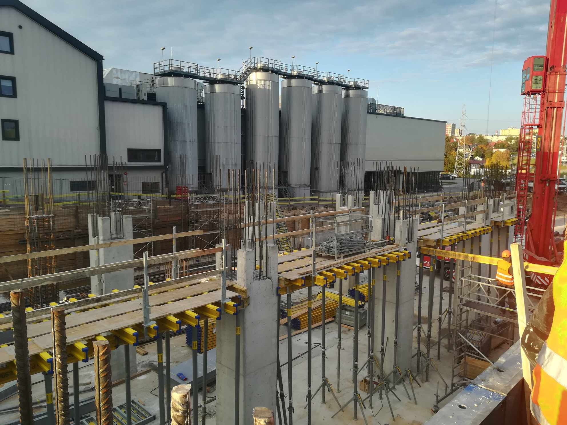 Wykop fundamentowy z betonowymi słupami i szalunkami, widoczne stalowe zbrojenia i dźwig w tle, obok budynek przemysłowy z metalowymi silosami.