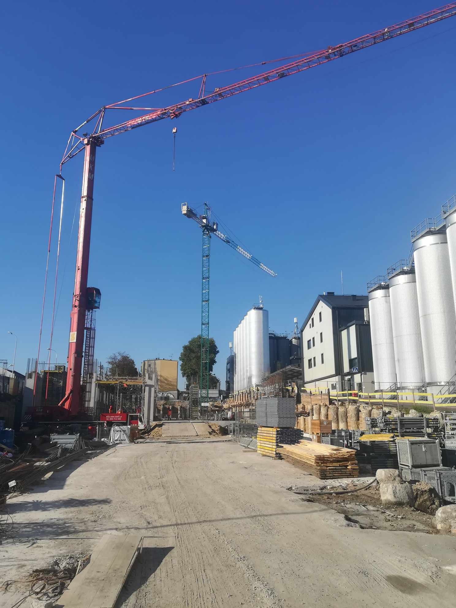 Plac budowy z dwoma żurawiami wieżowymi na tle błękitnego nieba i zabudowań przemysłowych, widoczne materiały budowlane i elementy konstrukcyjne.