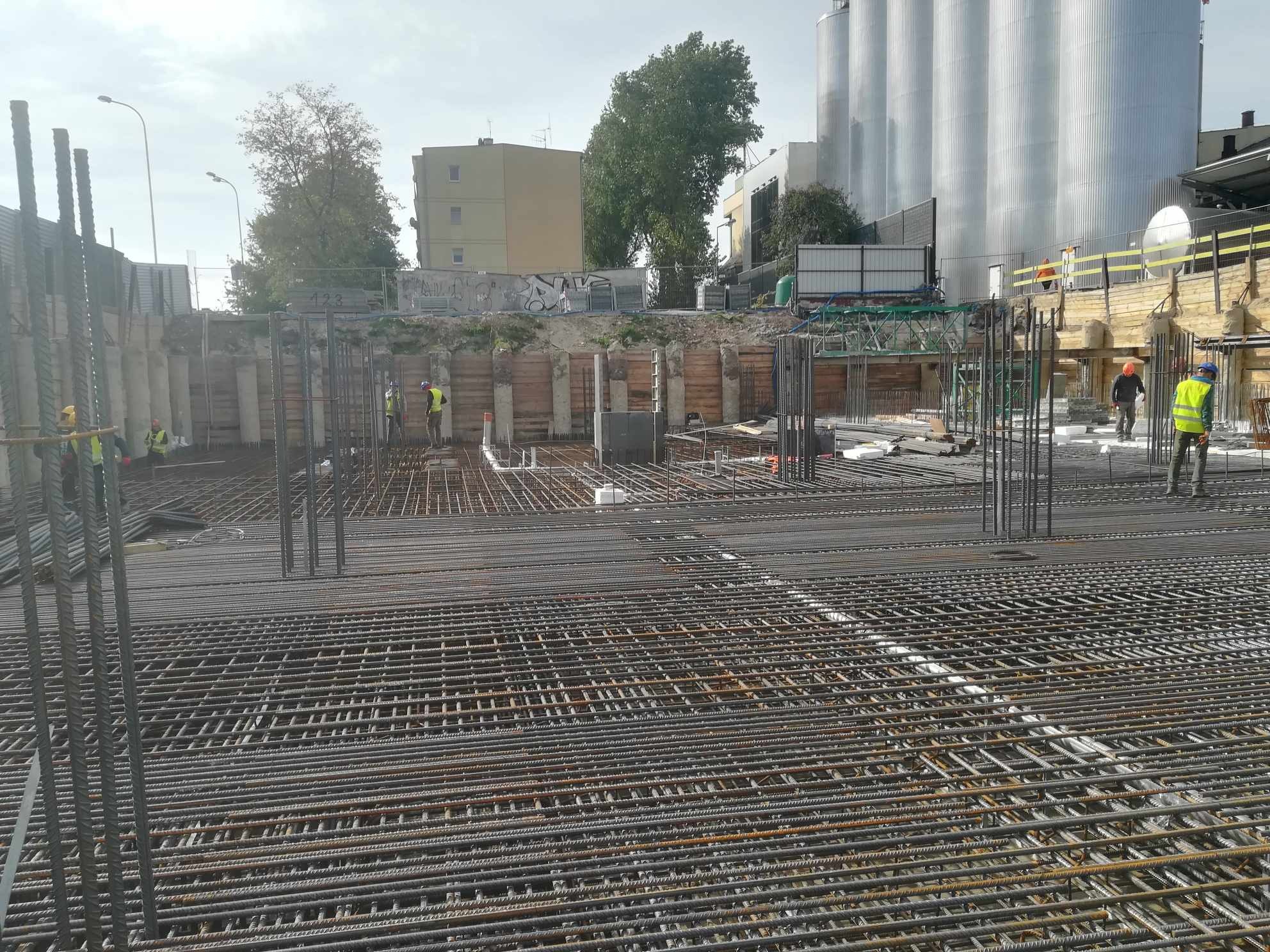 Zbrojenie fundamentu z widocznymi prętami stalowymi, szalunkiem i pracownikami w odblaskowych kamizelkach na placu budowy, w tle budynki i silosy.