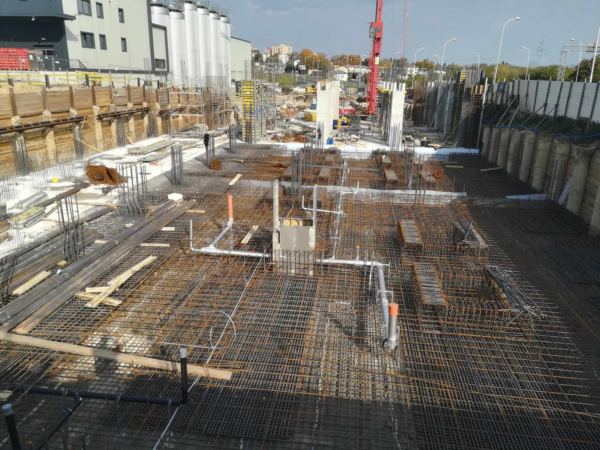 Zbrojenie płyty fundamentowej z instalacją kanalizacyjną, widok z góry na plac budowy z żurawiem i budynkami w tle.