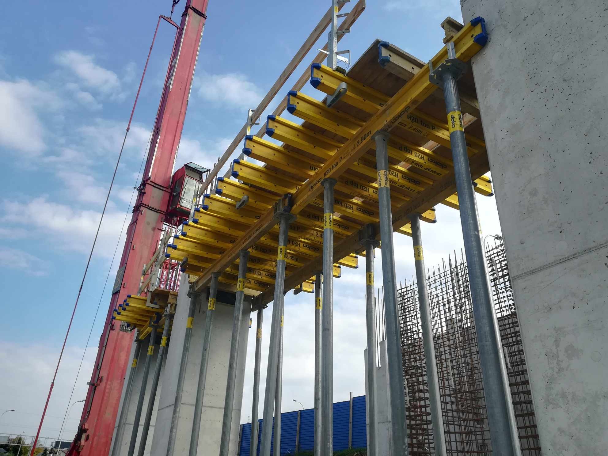 Konstrukcja szalunkowa z żółtymi belkami i stalowymi podporami, widoczna w trakcie budowy betonowego elementu, z żurawiem w tle i zbrojeniem w pobliżu.