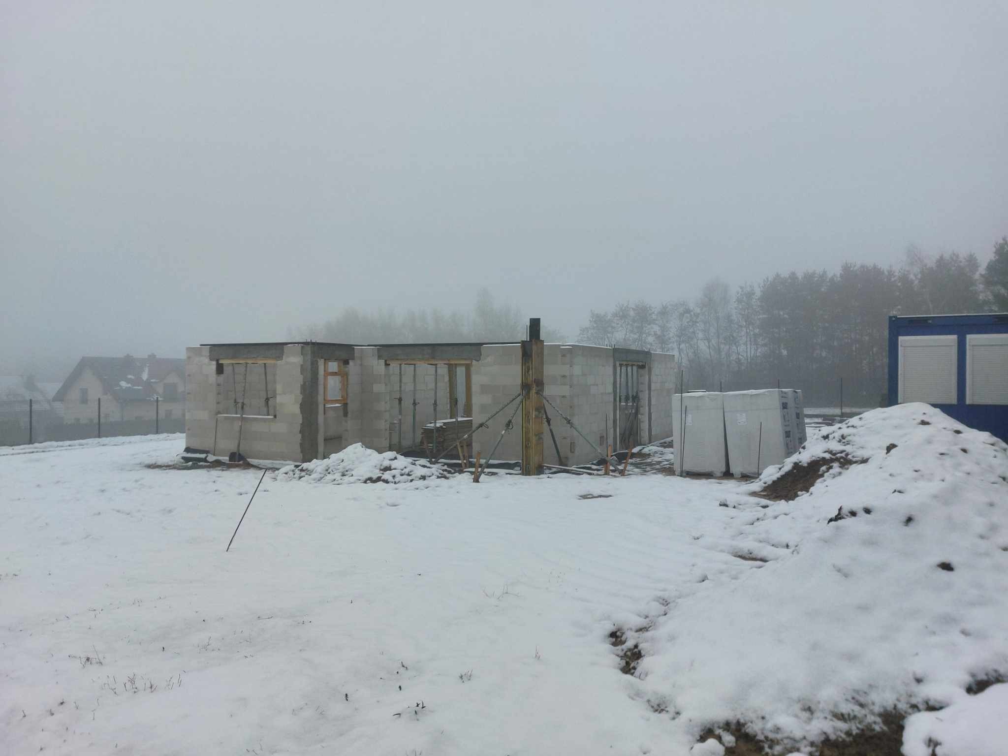 Niedokończony dom murowany z bloczków w zimowej scenerii, widoczne drewniane stemple podtrzymujące strop, a w tle zarys lasu i zabudowań w gęstej mgle.