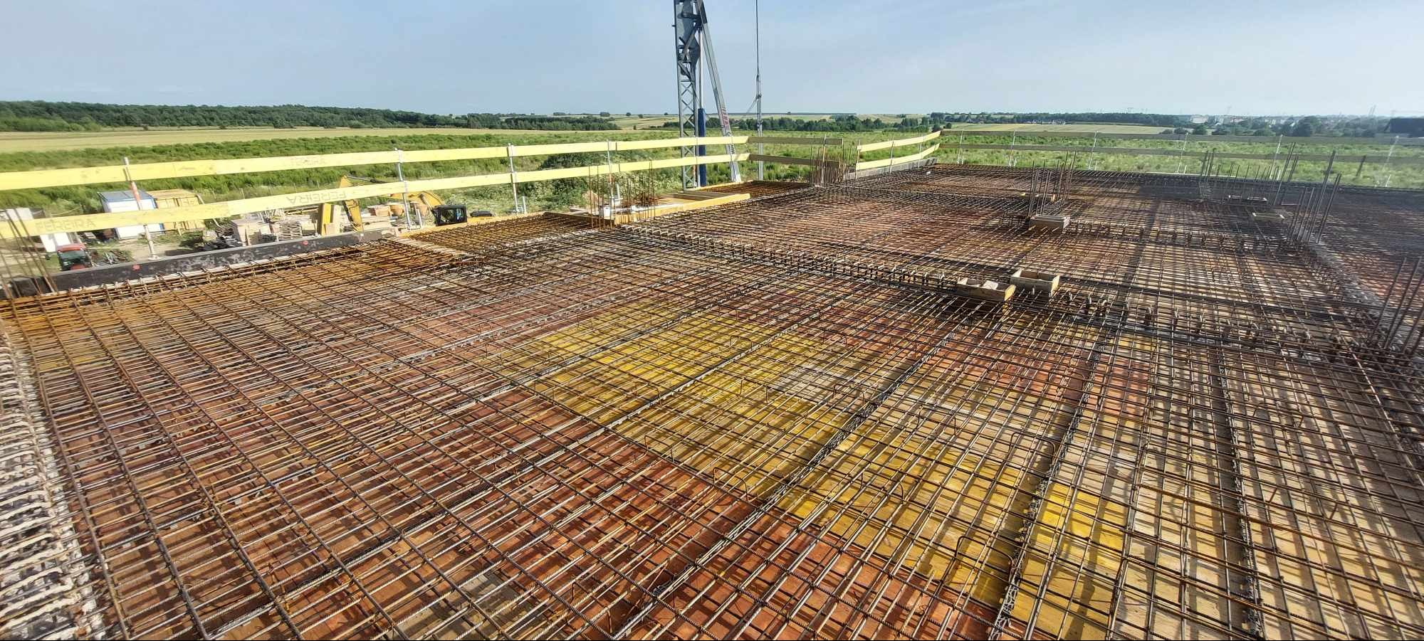 Zbrojenie płyty fundamentowej z widocznymi prętami stalowymi ułożonymi w siatkę na żółtych deskach szalunkowych, w tle krajobraz z zielonymi polami i lasem oraz żuraw budowlany.