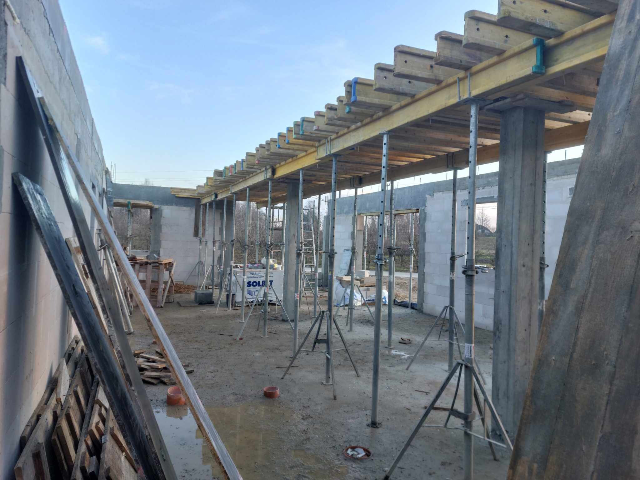 Wnętrze surowego domu w trakcie budowy, widoczne betonowe słupy podtrzymujące drewniane szalunki stropu, stalowe podpory, materiały budowlane w tle i mokra posadzka.