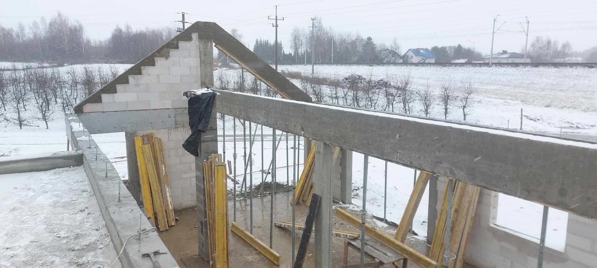 Surowy stan domu w trakcie budowy, widoczne ściany z bloczków, betonowy wieniec i drewniane stemple podpierające, zimowy krajobraz w tle.
