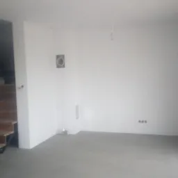 Surowe wnętrze po remoncie: drewniane schody z czarną konstrukcją, świeżo pomalowane białe ściany z widocznymi gniazdkami elektrycznymi i wentylacją, szara podłoga, okno po prawej stronie.