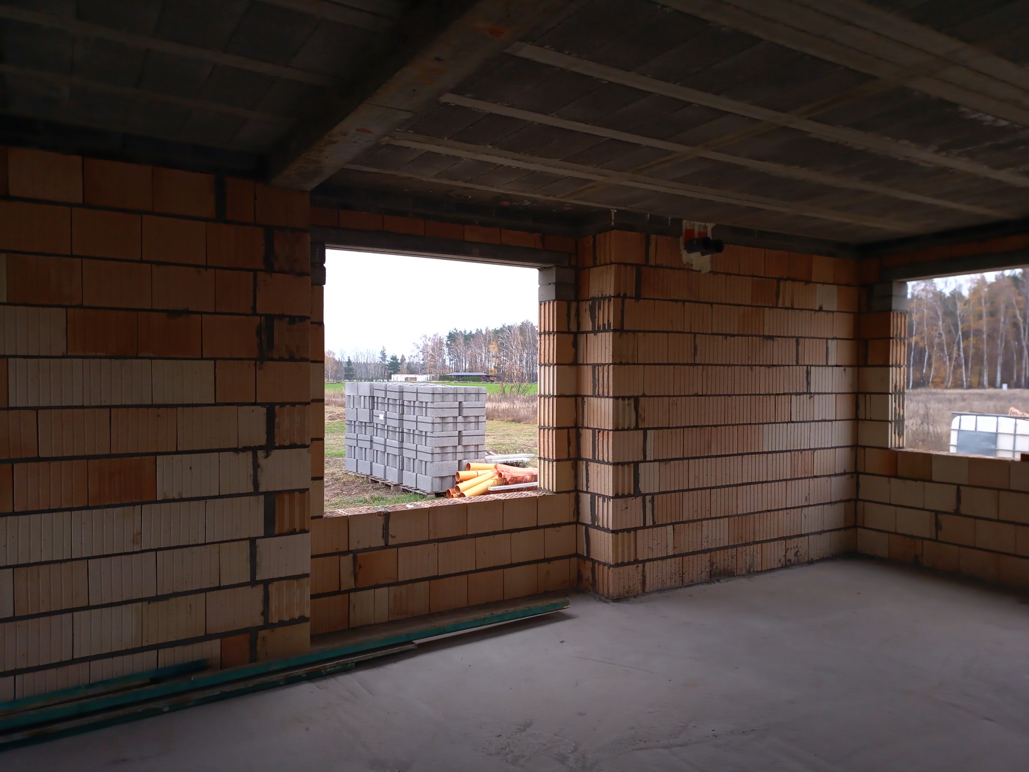 Wnętrze surowego budynku w trakcie budowy z widocznymi ścianami z cegieł, betonowym stropem i oknami wychodzącymi na stos materiałów budowlanych na zewnątrz.