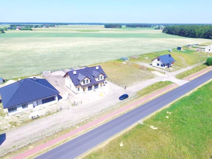 Panoramiczny widok z lotu ptaka na dwa domy jednorodzinne z ciemnymi dachami, otoczone polami i drogą asfaltową w słoneczny dzień.