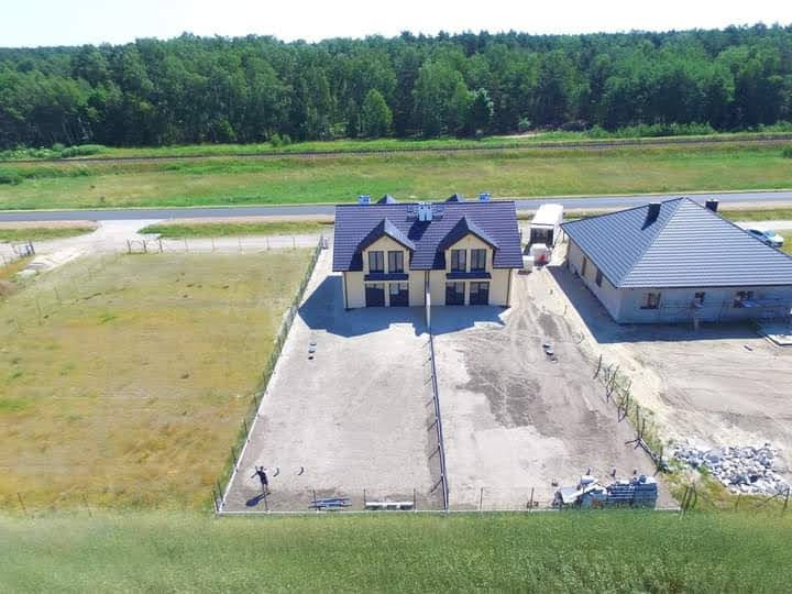Widok z lotu ptaka na nowo wybudowany dom dwurodzinny z ciemnym dachem i jasną elewacją, obok drugi budynek w trakcie wykończenia, teren wokół wyrównany, częściowo ogrodzony, w tle las i linia...