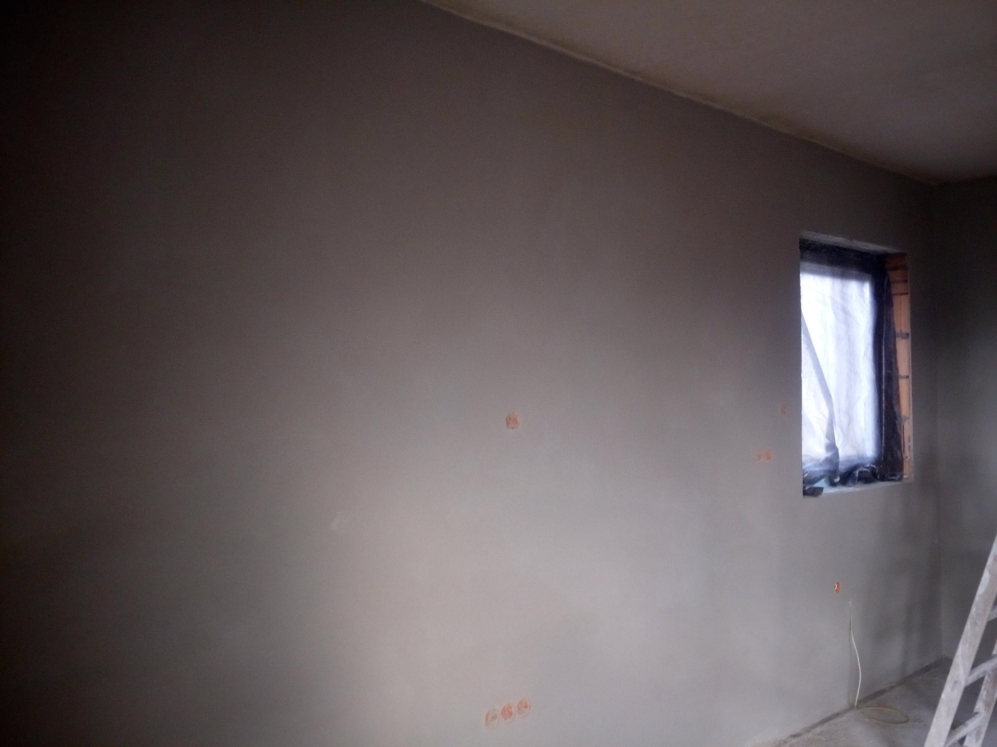 Ściana po nałożeniu gładzi szpachlowej, z widocznymi puszkami elektrycznymi i oknem zabezpieczonym folią malarską, drabina oparta o ścianę z prawej strony.