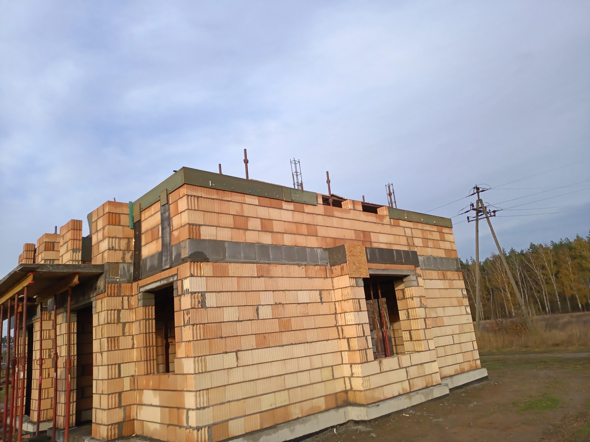 Surowy stan budynku jednorodzinnego z pustaków ceramicznych, widoczne zbrojenia, betonowe nadproża, szalunki i podpory stropu, w tle słup wysokiego napięcia i las.