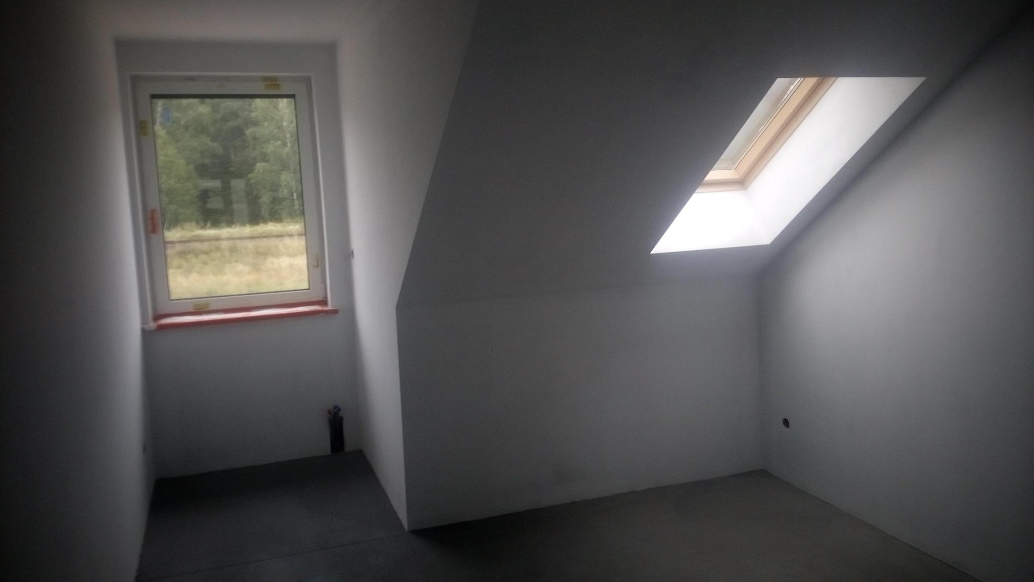 Poddasze po remoncie z oknem dachowym i oknem pionowym z widokiem na zewnątrz, świeżo pomalowane ściany na szaro, widoczny fragment czarnej rury przy ścianie.