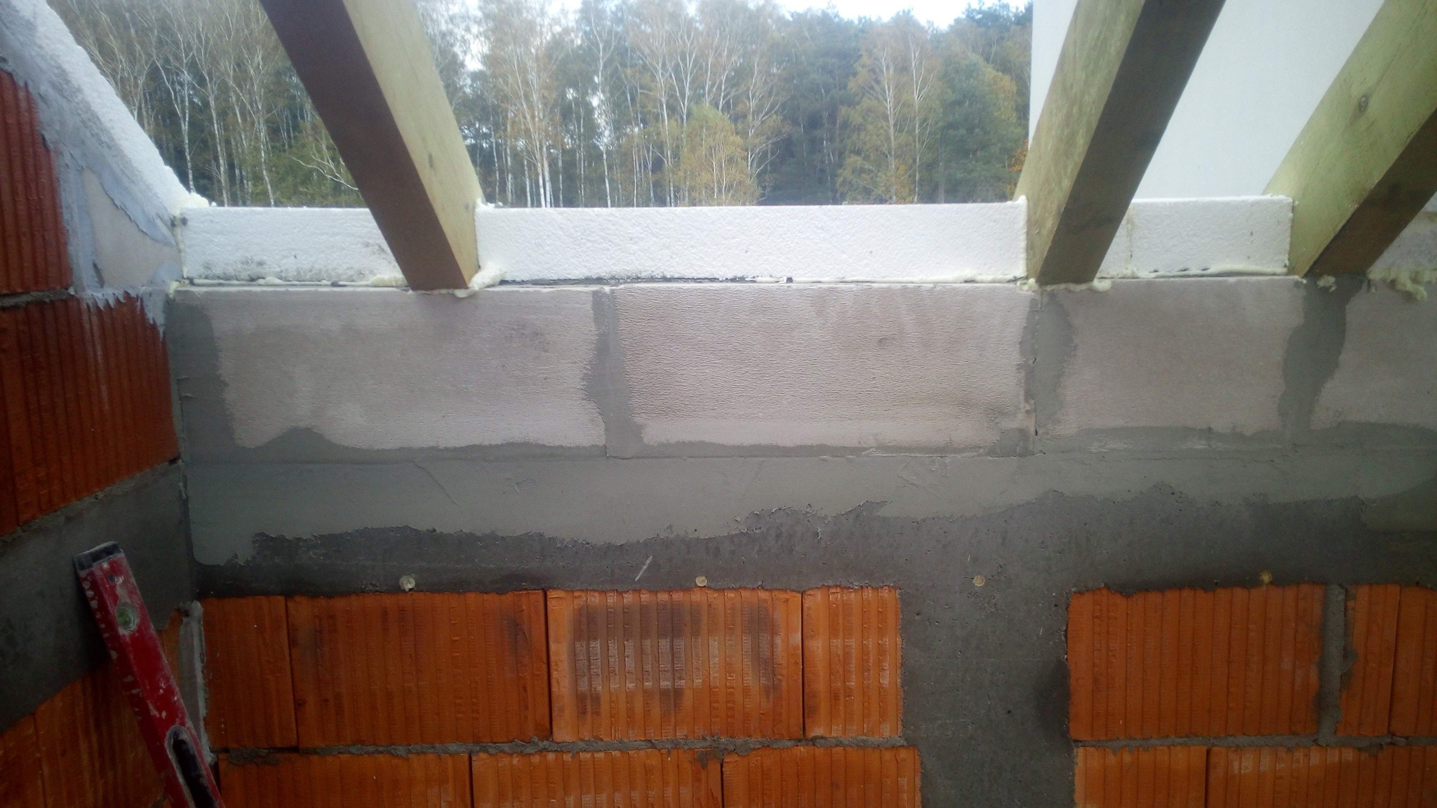 Wznoszenie muru z cegły ceramicznej, z widocznym poziomem murarskim, izolacją termiczną i drewnianymi elementami konstrukcyjnymi, na tle drzew