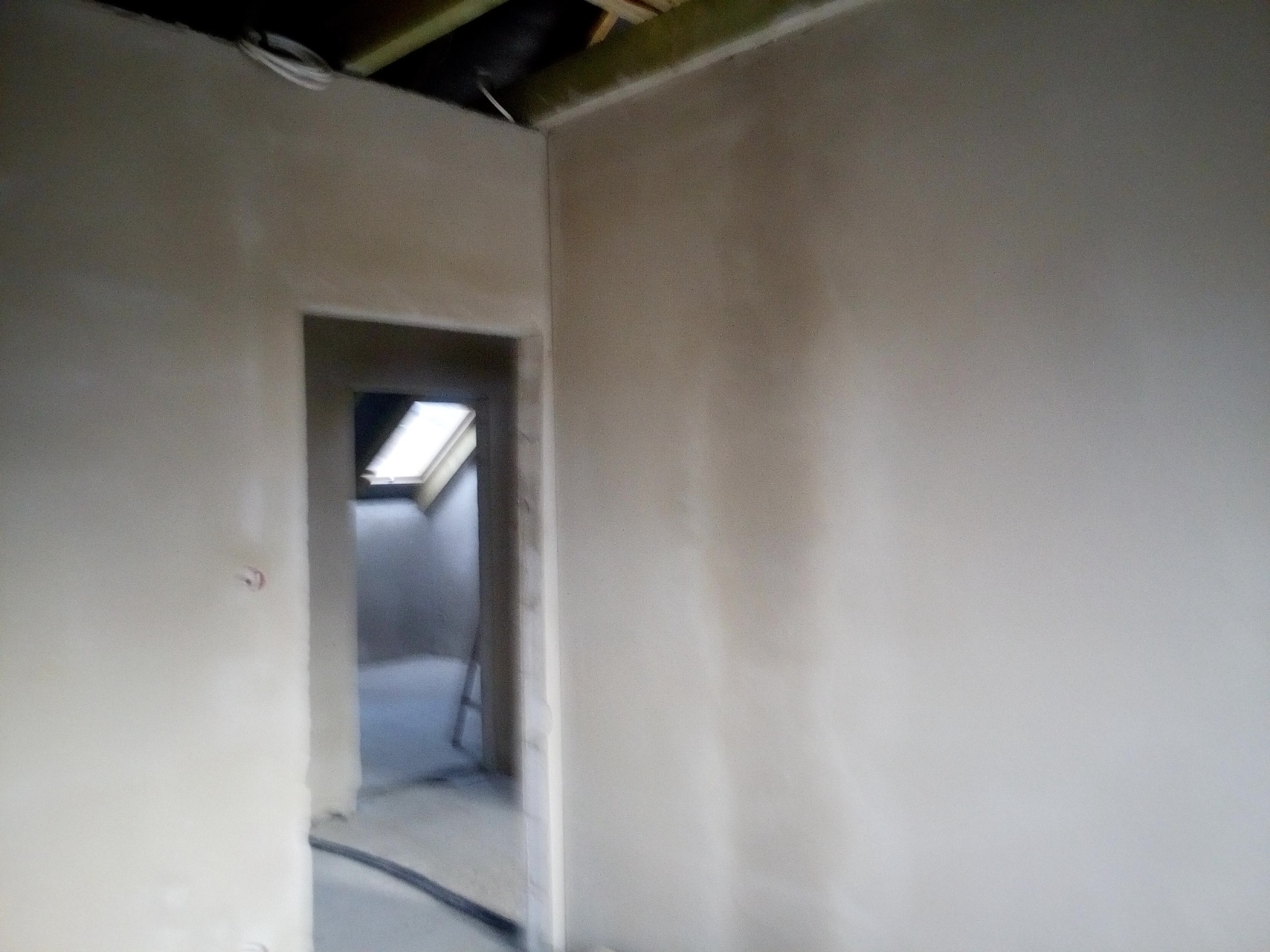 Wnętrze w trakcie gładzenia ścian, widoczne szpachlowane ściany, otwór drzwiowy prowadzący do pomieszczenia z oknem dachowym oraz fragment instalacji elektrycznej pod sufitem.