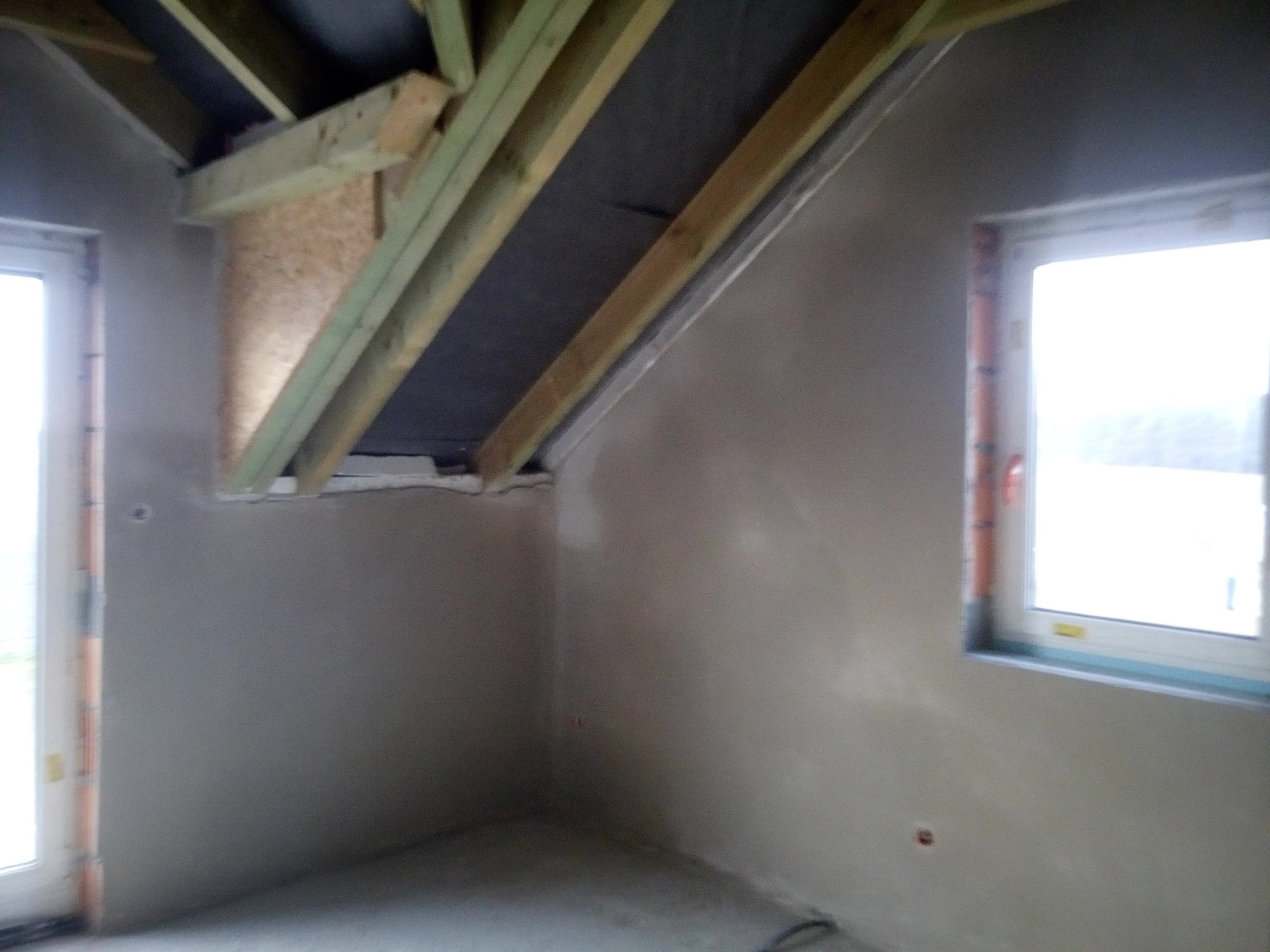 Surowe wnętrze poddasza z oknem i widoczną konstrukcją dachu przed wykończeniem ścian.