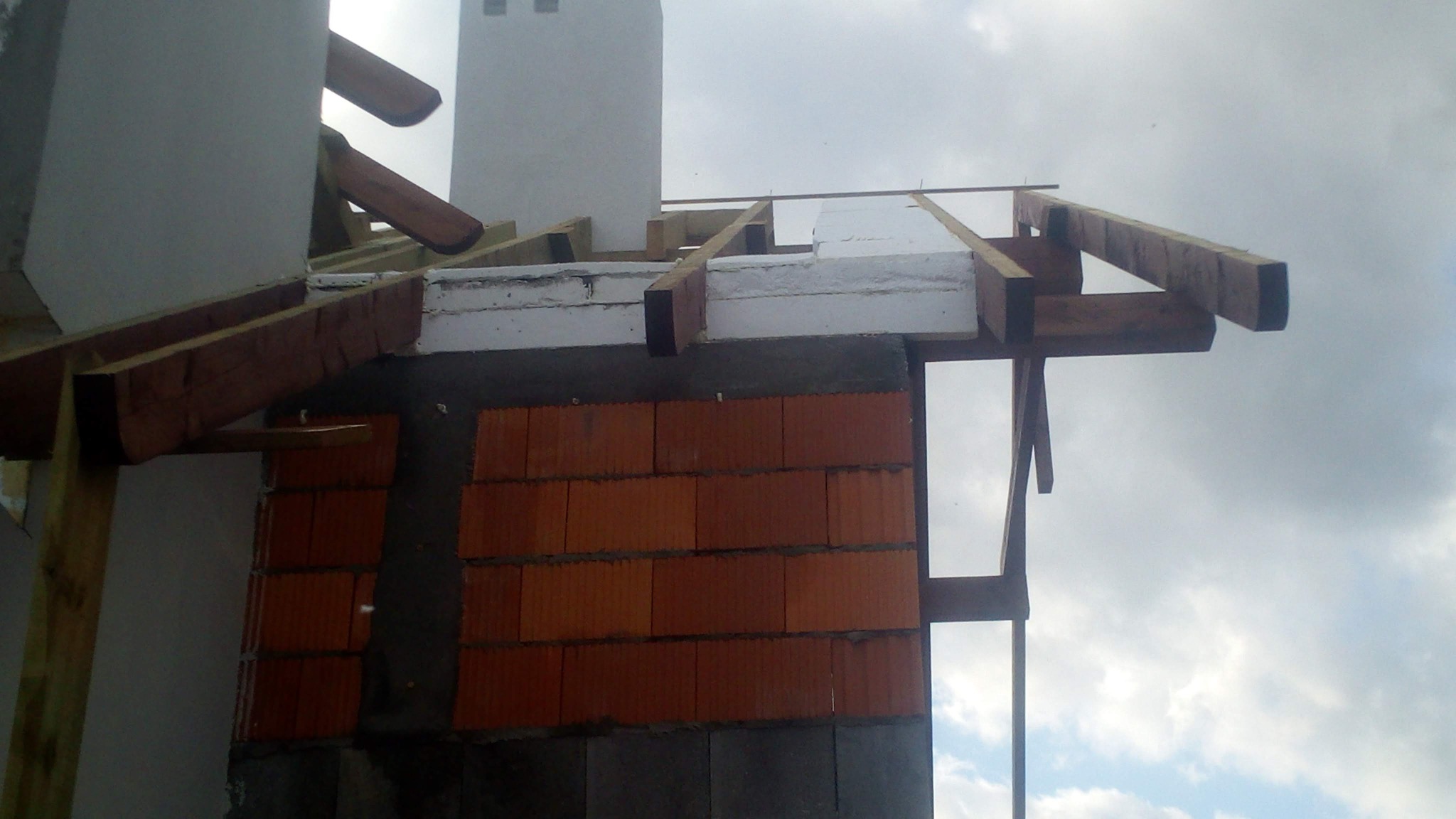 Wznoszenie muru z czerwonej cegły z widoczną konstrukcją drewnianą i ociepleniem styropianowym, fragment komina w tle.