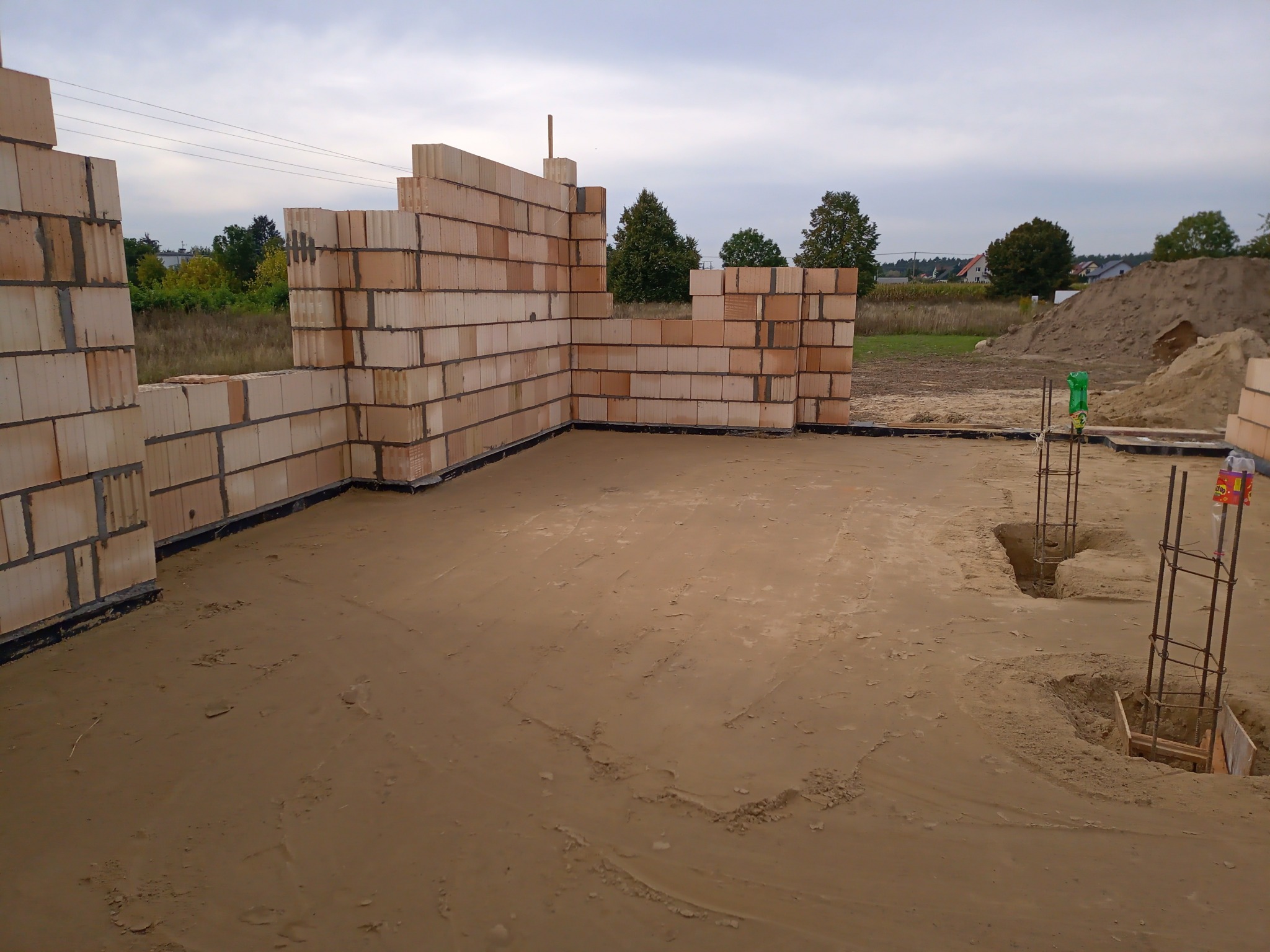 Wznoszone ściany z bloczków ceramicznych na fundamencie, widoczne zbrojenia w wykopach, w tle hałda piasku i zabudowa jednorodzinna.