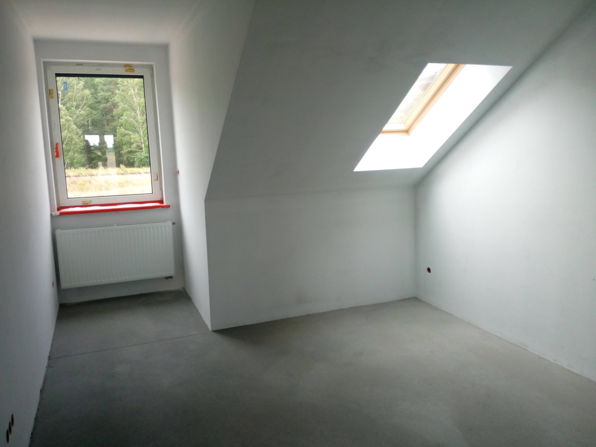 Surowe wnętrze poddasza z oknem dachowym i grzejnikiem, ściany pomalowane na biało, w tle widok na drzewa.