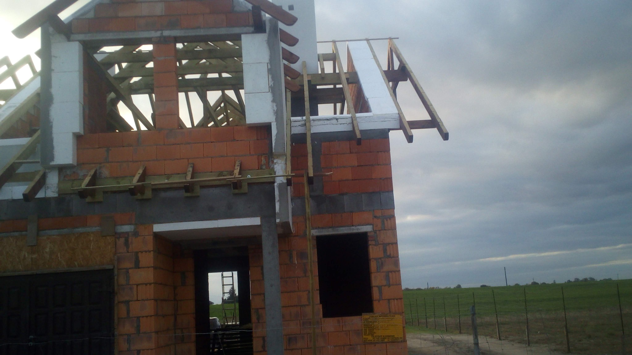 Surowy stan domu w trakcie budowy: czerwona cegła, drewniana konstrukcja dachu z izolacją, widoczne rusztowania i drabina, tło pochmurne niebo i zielone pole.