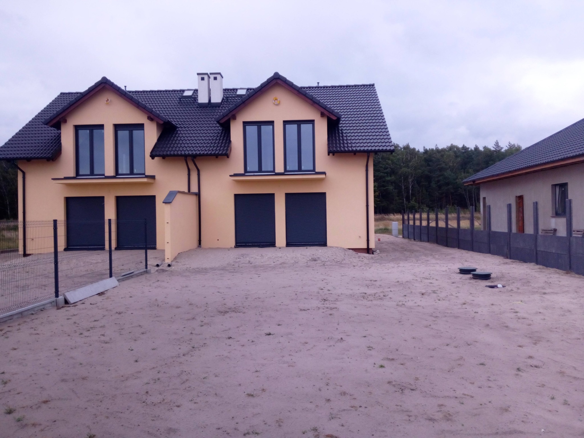 Dwa identyczne, nowo wybudowane domy bliźniacze z szarą dachówką i roletami w oknach garażowych, oddzielone od ulicy surowym ogrodzeniem z siatki, na niezagospodarowanej działce z piaskiem.