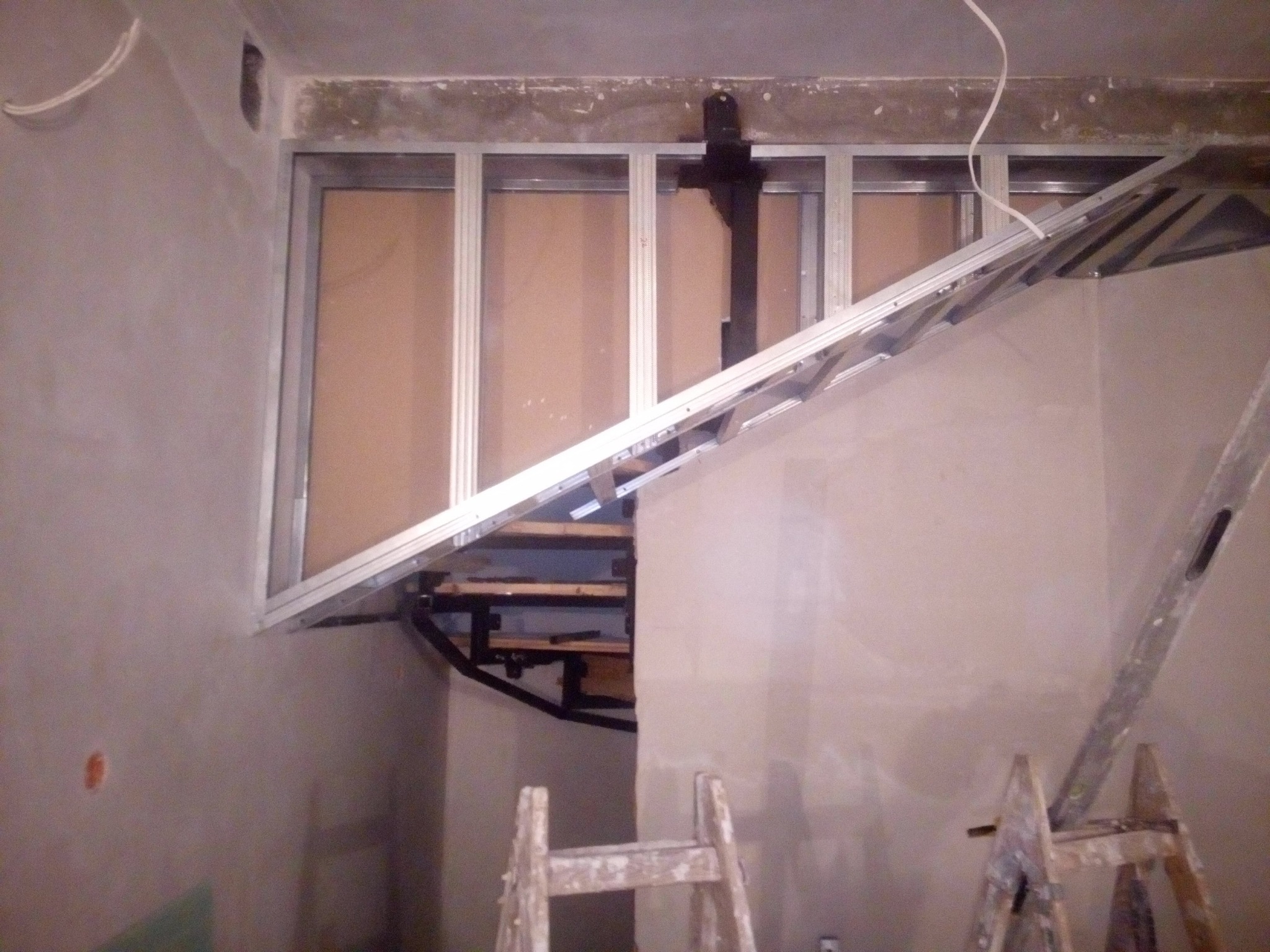 Metalowa konstrukcja sufitu podwieszanego w trakcie montażu, widoczne profile i drabina malarska w pomieszczeniu o surowych ścianach.