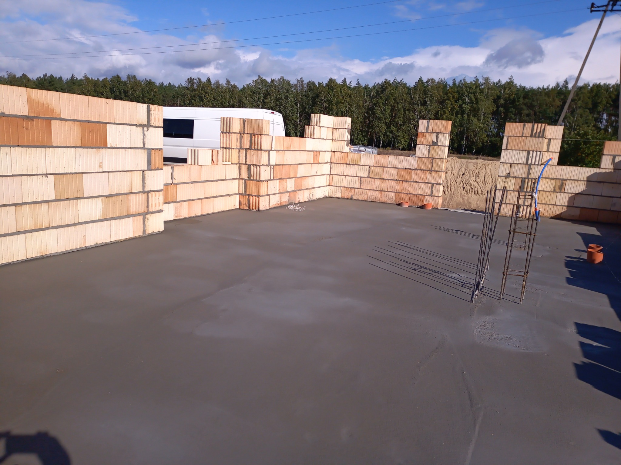 Wylany betonowy fundament pod budynek z częściowo wzniesionymi ścianami z bloczków ceramicznych, widoczny szary beton, zbrojenie i biały bus w tle.