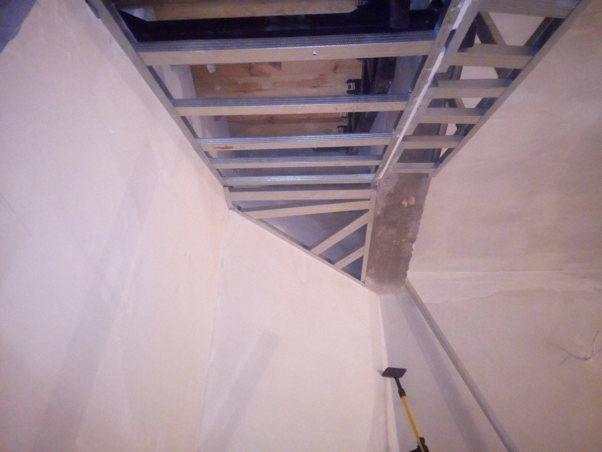 Sufit podwieszany z metalową konstrukcją w trakcie montażu, widoczne przewody i instalacje powyżej, ściany przygotowane do malowania, żółta rączka narzędzia w rogu.