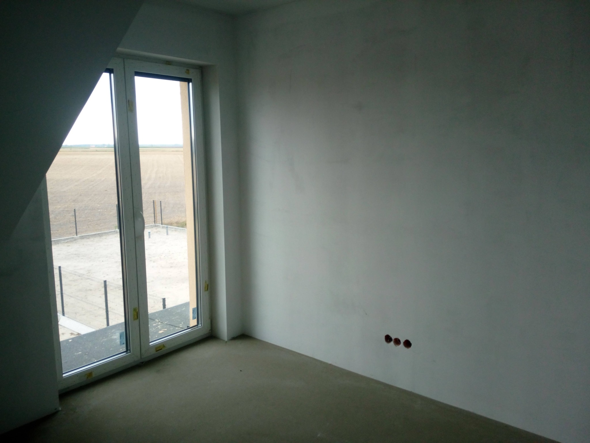 Surowy pokój na poddaszu z oknem wychodzącym na pole; ściany świeżo otynkowane, widoczne puszki elektryczne.