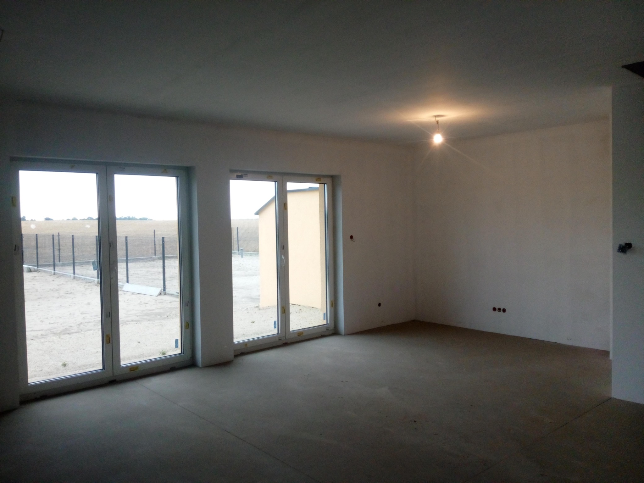 Surowe wnętrze nowego domu z betonową podłogą, widoczne okna tarasowe z widokiem na ogrodzenie i sąsiedni budynek. Na suficie wisi pojedyncza żarówka.