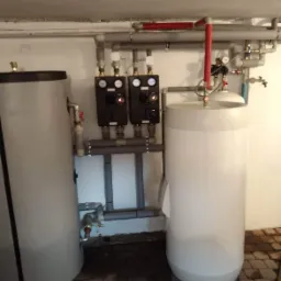 Dwa zbiorniki na wodę użytkowa z panelami sterowania i rurami w pomieszczeniu technicznym na podłodze z kostki brukowej.