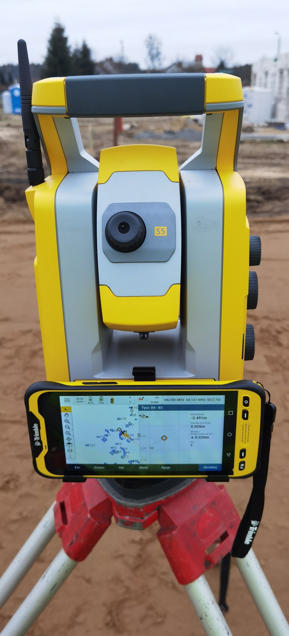 Żółto-szary tachimetr z zamocowanym smartfonem, wyświetlającym mapę geodezyjną, na tle placu budowy z widocznymi fundamentami i elementami konstrukcyjnymi.