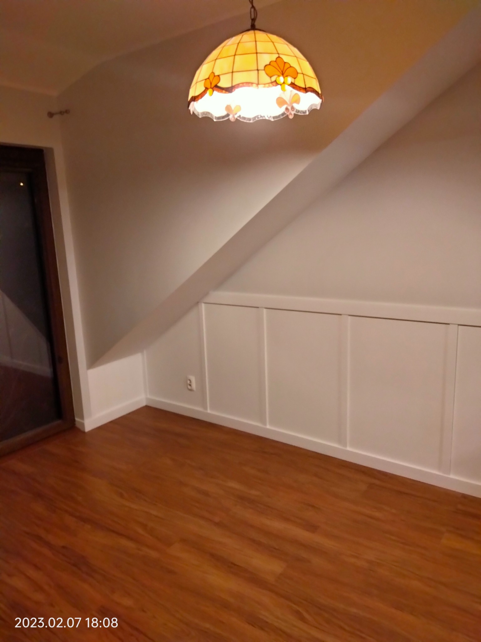 Poddasze z drewnianą podłogą, białą boazerią panelową pod skosem i ozdobną lampą witrażową w stylu Tiffany.