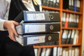 Stos trzech czarnych segregatorów z etykietami trzymanych przez osobę w białej koszuli, z regałem w tle. Dokumenty firmowe.