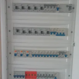 Otwarta, biała skrzynka rozdzielcza z czterema rzędami bezpieczników i wyłączników różnicowoprądowych różnych producentów, widoczna tabliczka znamionowa ETI w prawym górnym rogu.