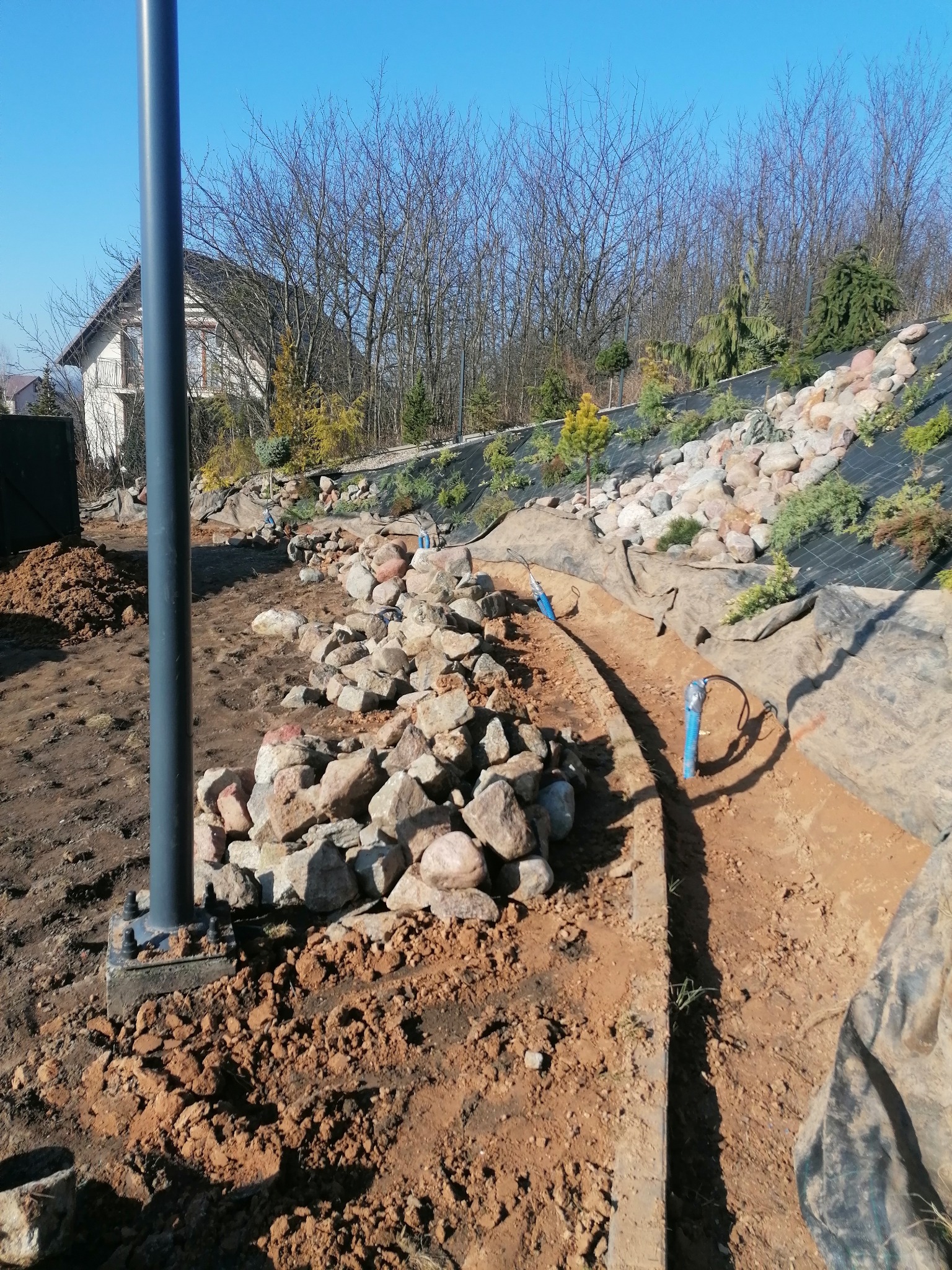 Budowa skalniaka w ogrodzie z widocznym systemem nawadniania, kamieniami i młodymi roślinami na tle czarnej agrowłókniny, w tle dom jednorodzinny.