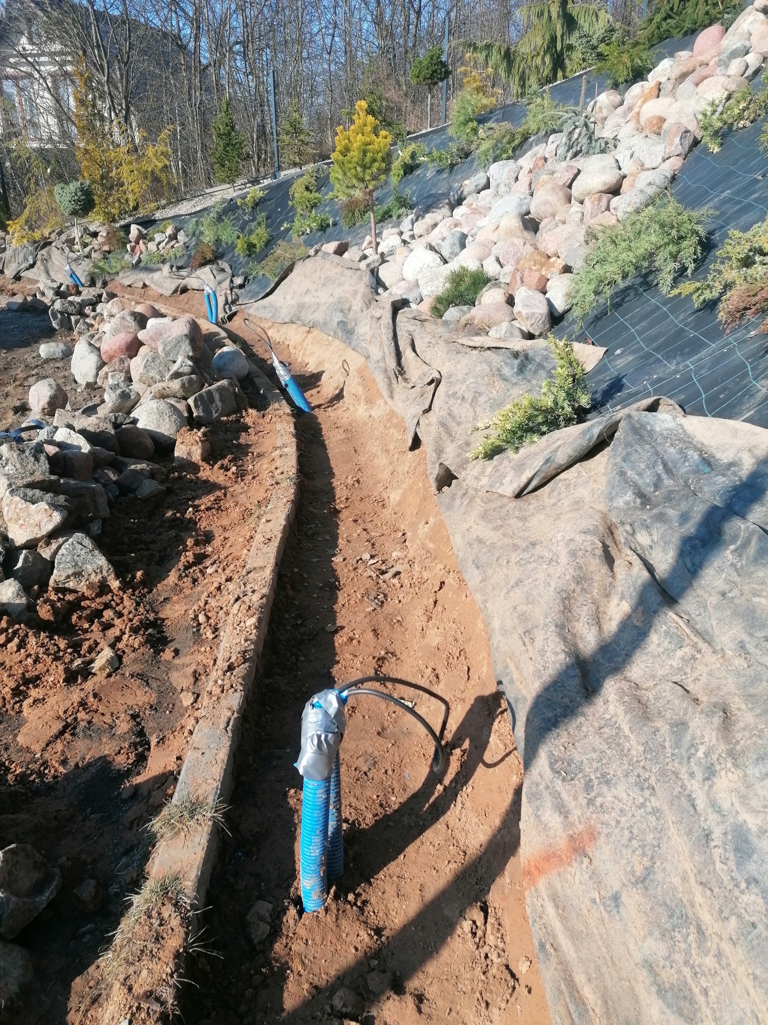 Montaż systemu nawadniania w Tucholi: Rów w ziemi z widocznymi niebieskimi rurami, na zboczu kamienie i rośliny zabezpieczone agrowłókniną.