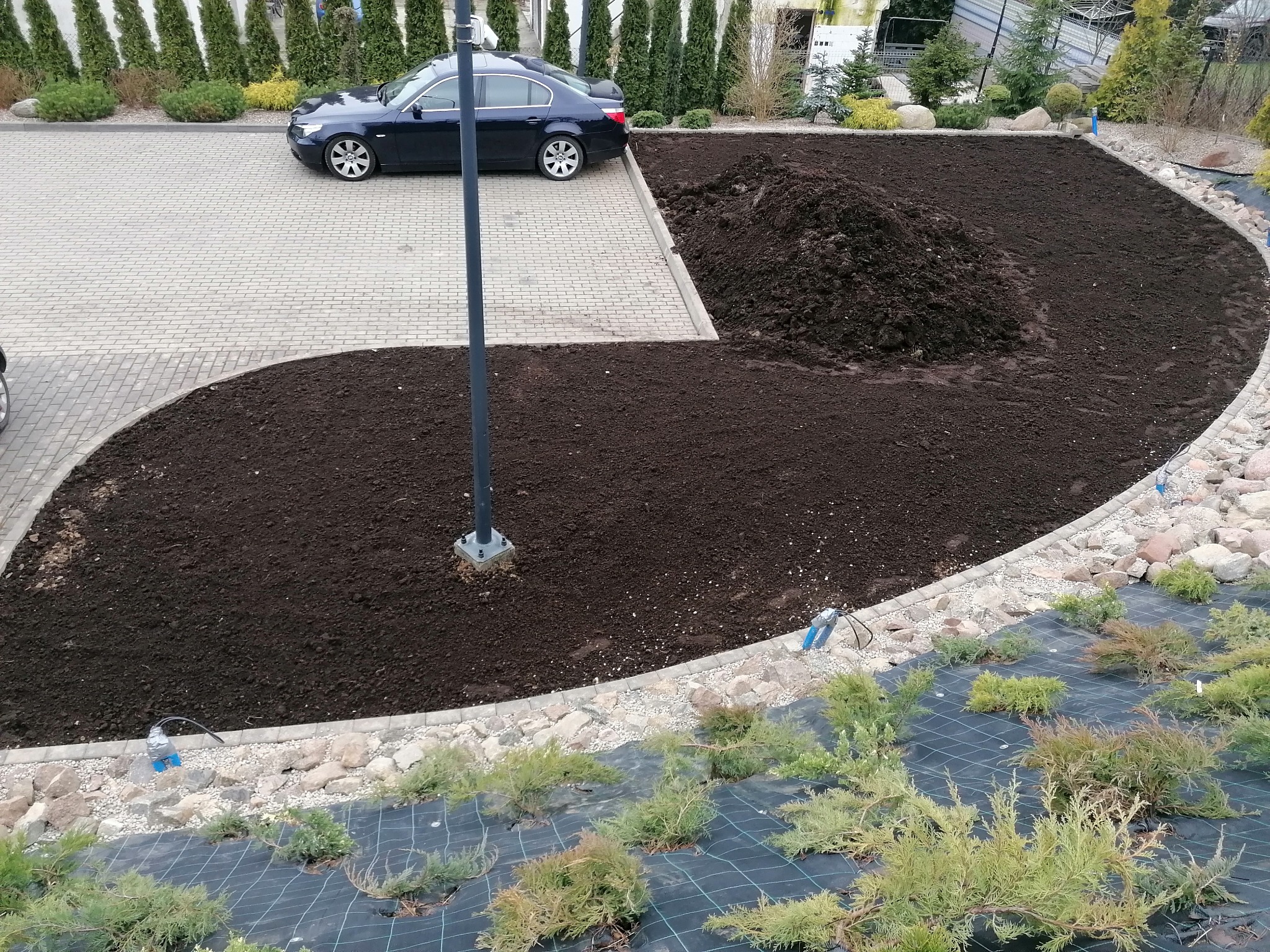 Przygotowanie terenu pod trawnik z rolki: wyrównana ziemia, czarna agrowłóknina i system nawadniania w ogrodzie przed domem z podjazdem i zaparkowanym samochodem.