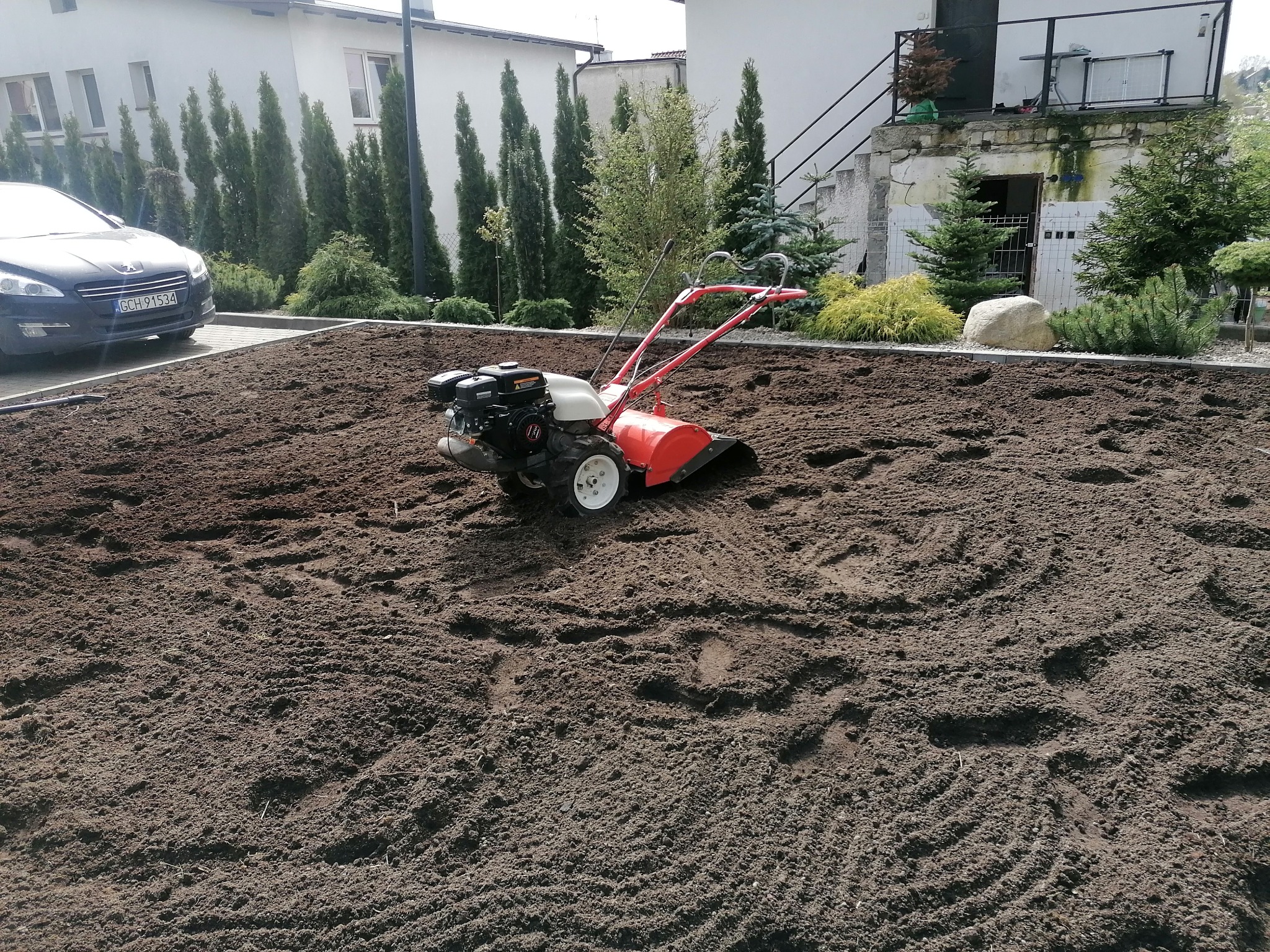 Gleba świeżo przekopana glebogryzarką w ogrodzie przydomowym, widoczne ślady pracy maszyny, w tle budynek mieszkalny i zaparkowany samochód.