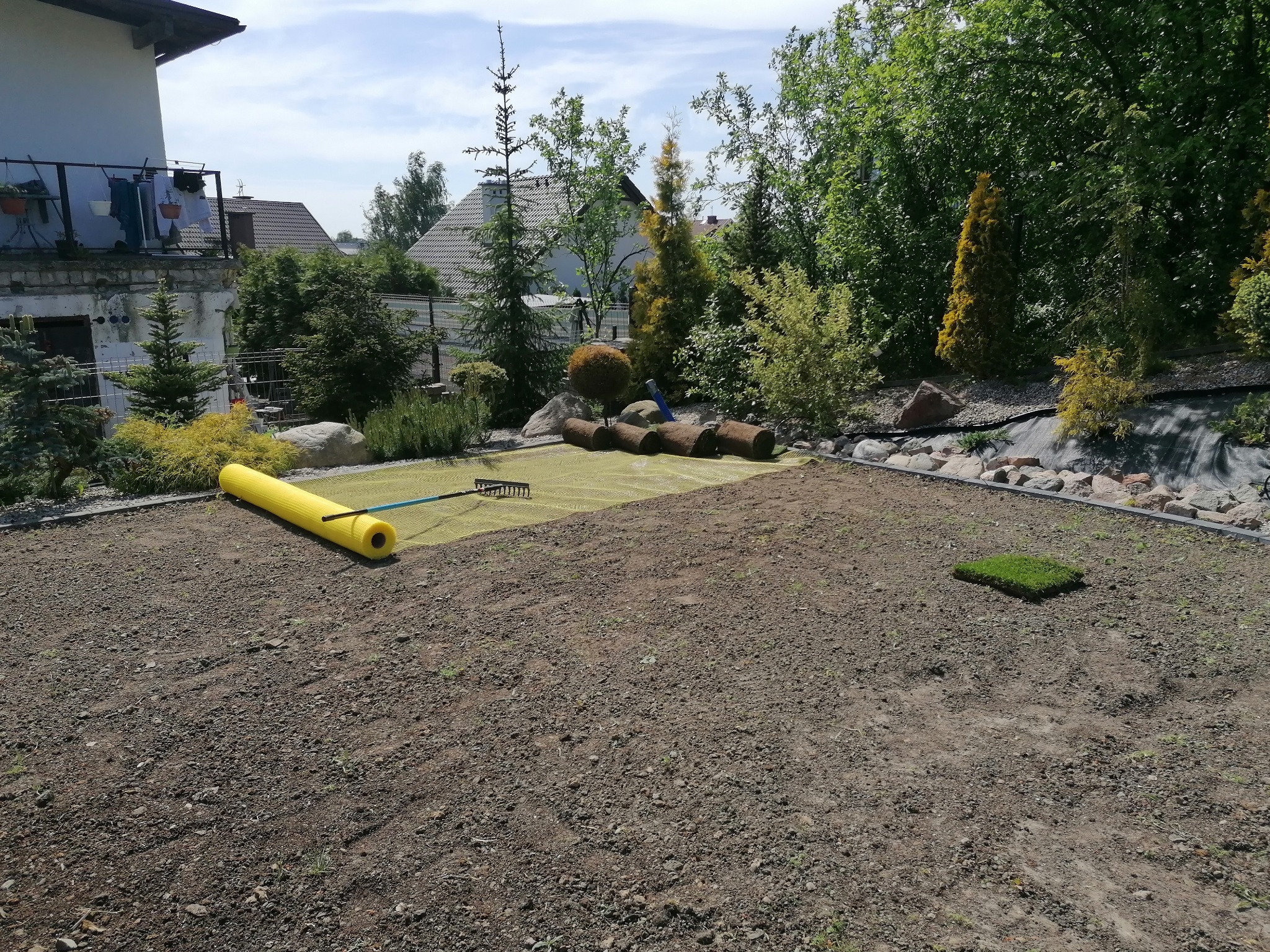Przygotowanie terenu pod trawnik z rolki: rozłożona siatka, rolki trawy i grabie na tle domu z balkonem.