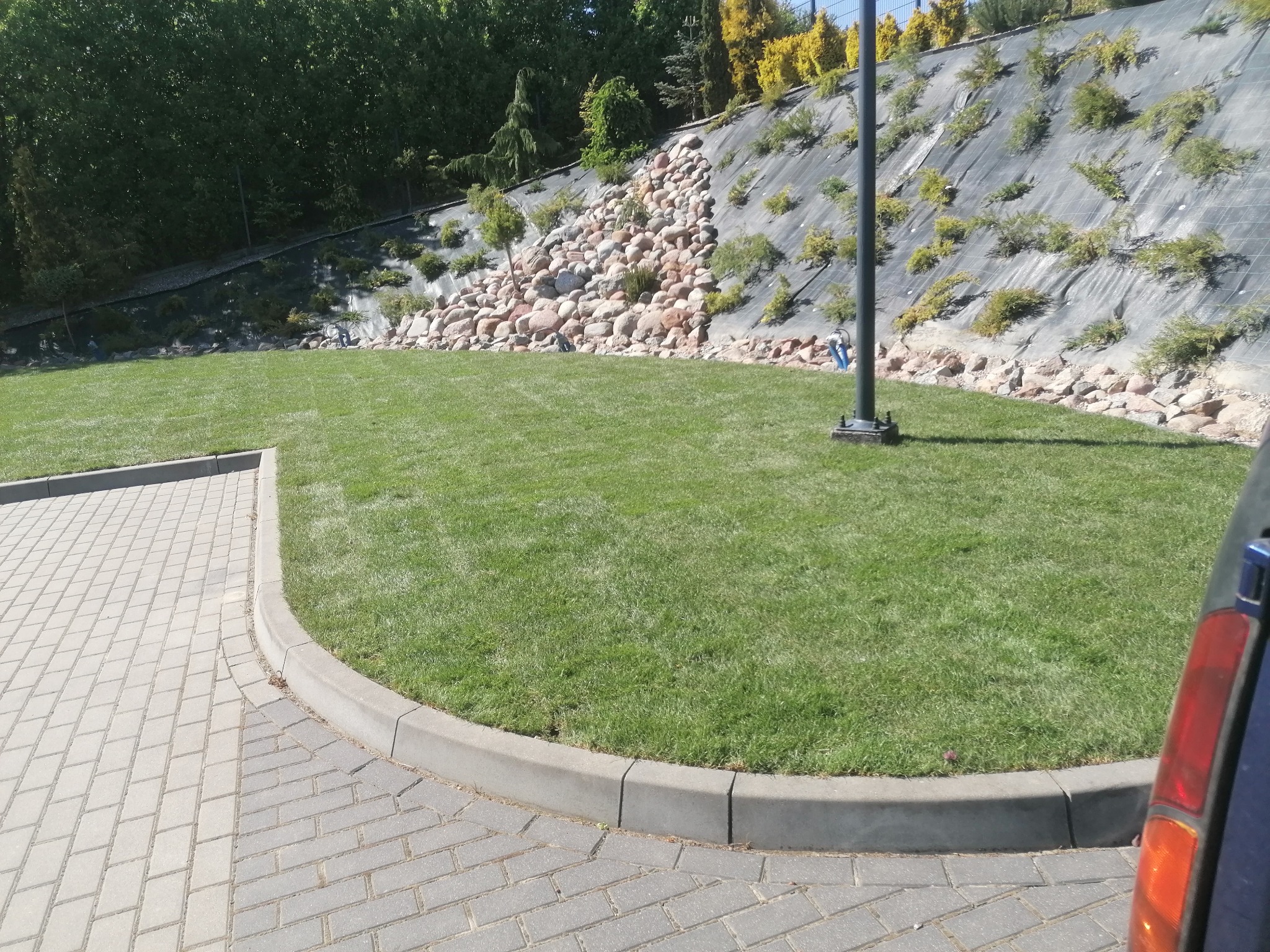 Starannie utrzymany trawnik przylegający do kostki brukowej, z dekoracyjnym skalniakiem na zboczu w tle.