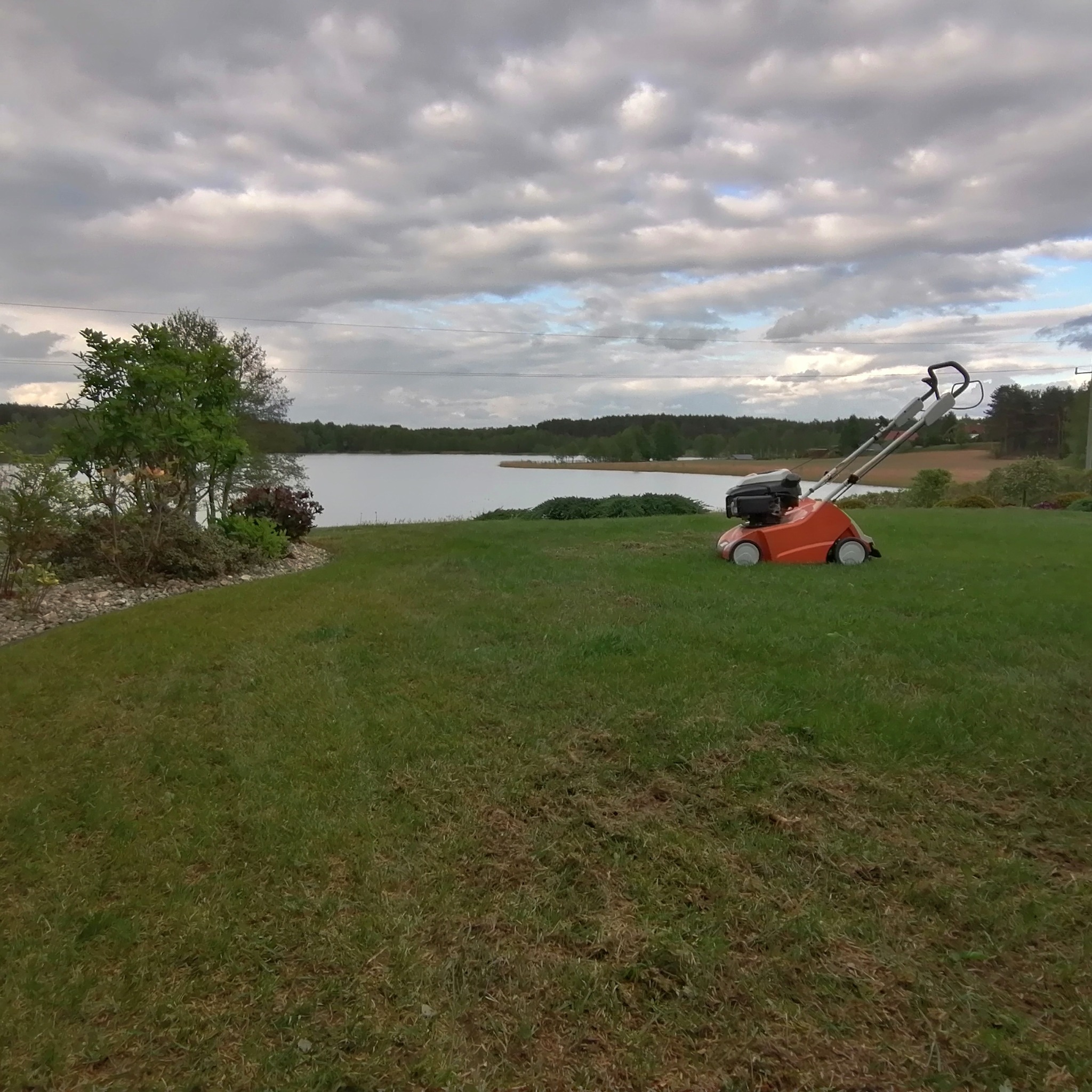 Pomarańczowa kosiarka spalinowa stoi na trawniku nad jeziorem pod pochmurnym niebem, widoczne ślady koszenia.