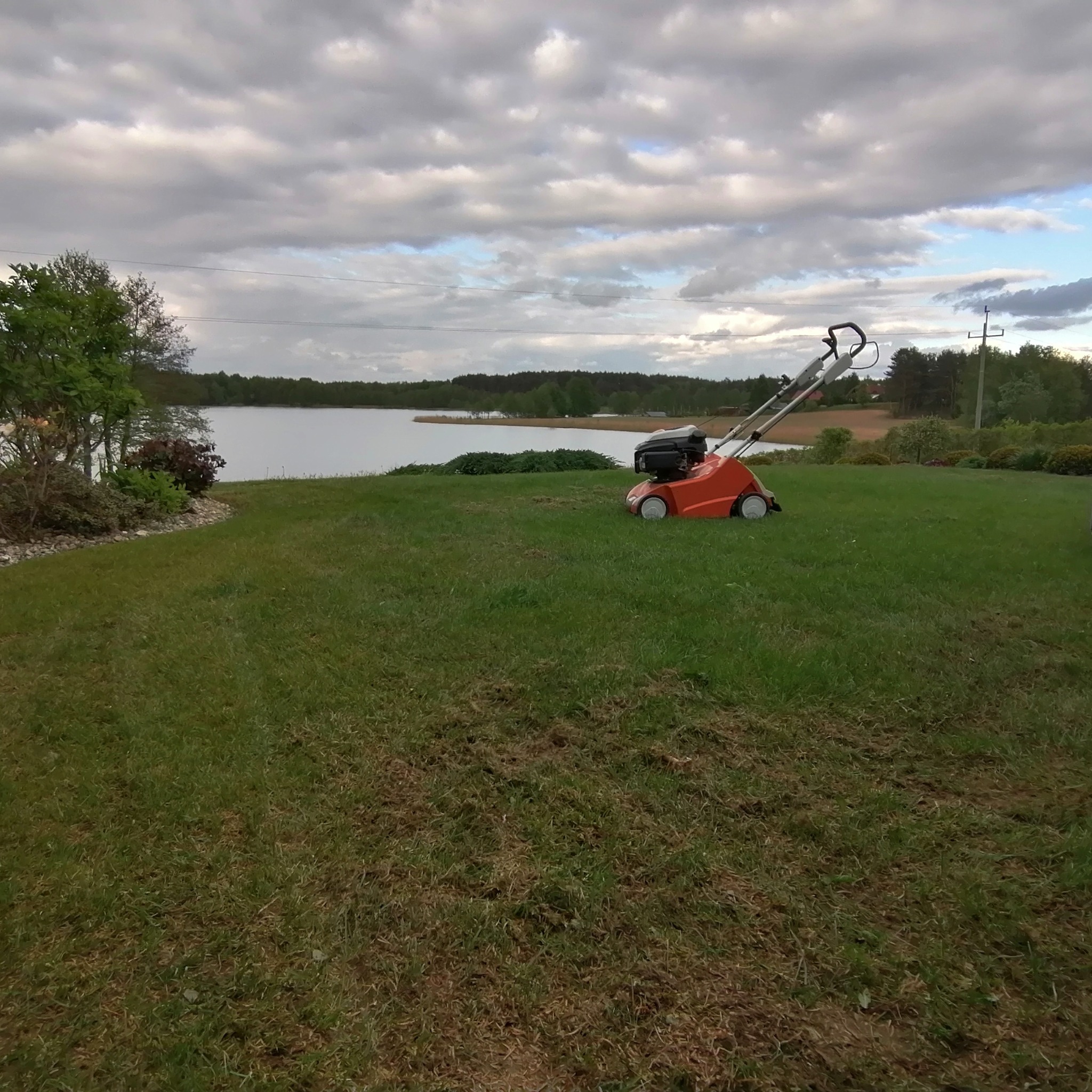 Pomarańczowa kosiarka spalinowa na trawniku nad brzegiem jeziora, widoczne ślady po koszeniu.