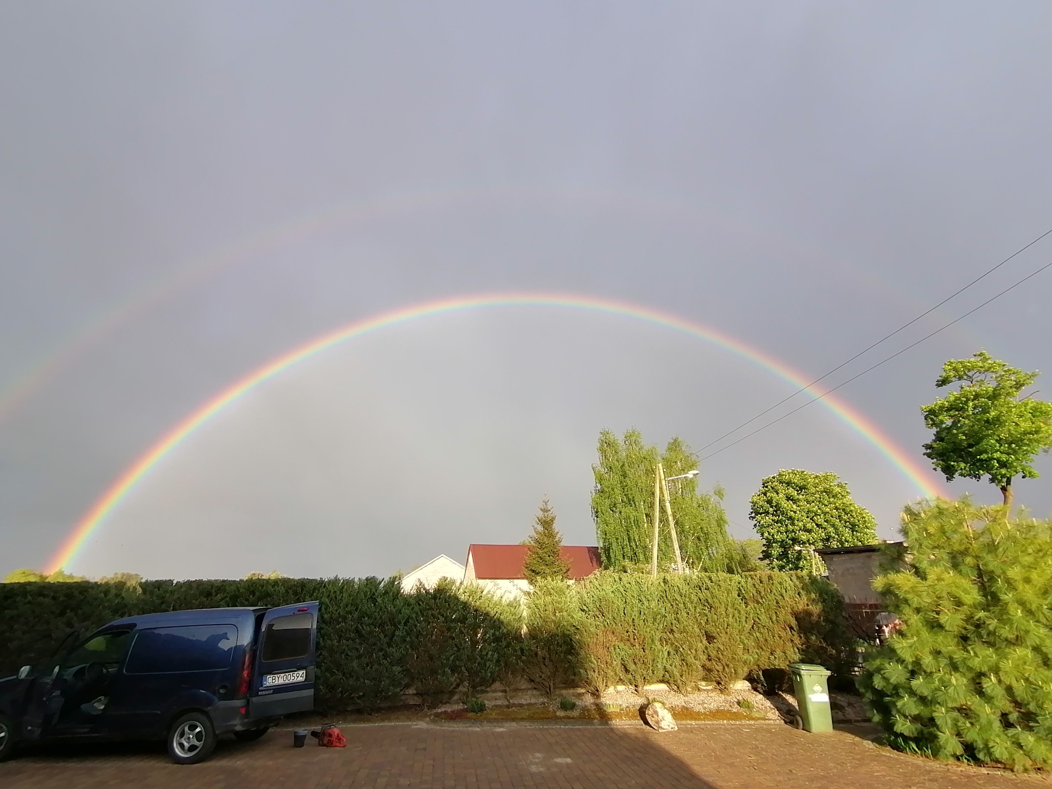 Podwójna tęcza nad zadbanym żywopłotem, z zaparkowanym niebieskim vanem z otwartymi drzwiami bagażnika, sugerującym przerwę w pracy ogrodniczej.